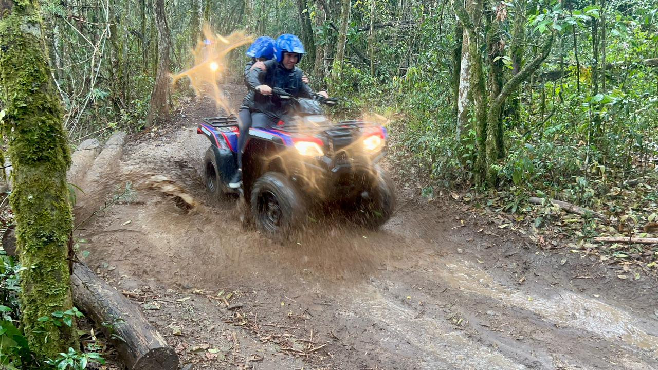 Dois passageiros de capacete azul atravessando barro na trilha.